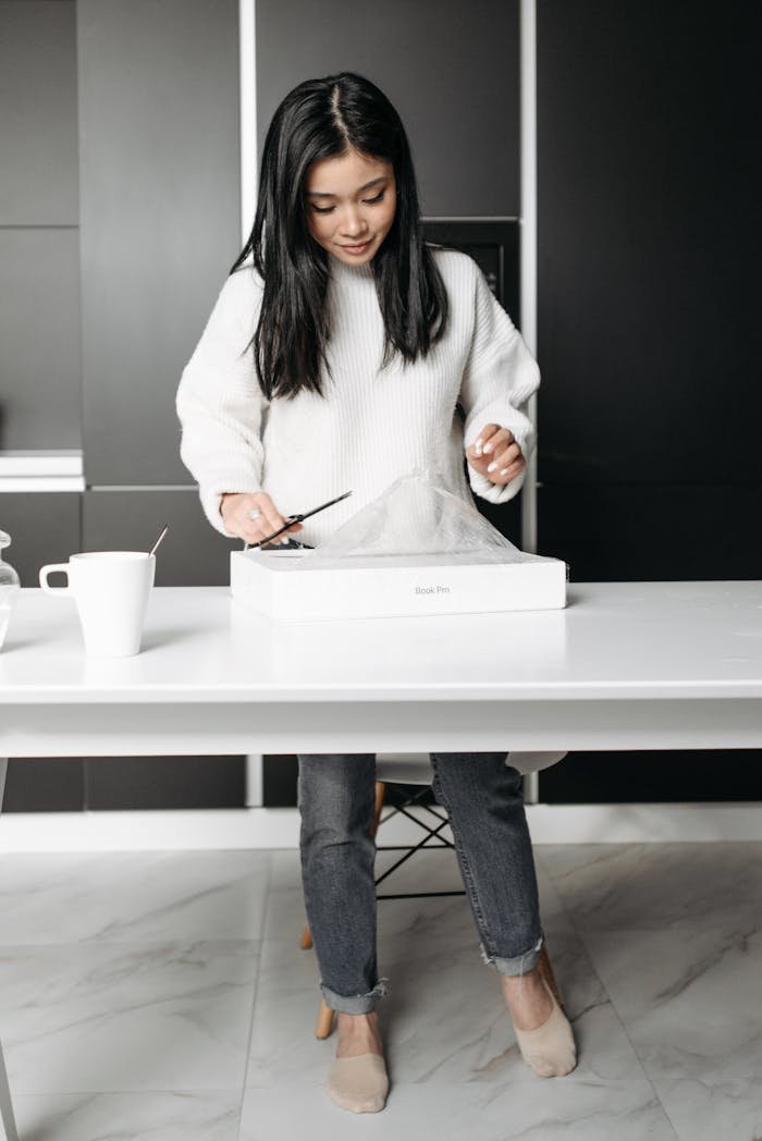 Asian woman opens new laptop in modern kitchen, showcasing excitement and anticipation.